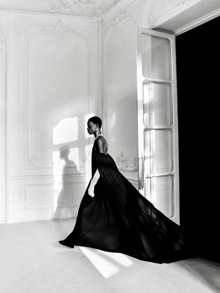 Чёрно-белый студийный кадр: модель в длинном тёмном платье идёт по комнате с высокими окнами, классический художественный образ коллекции.