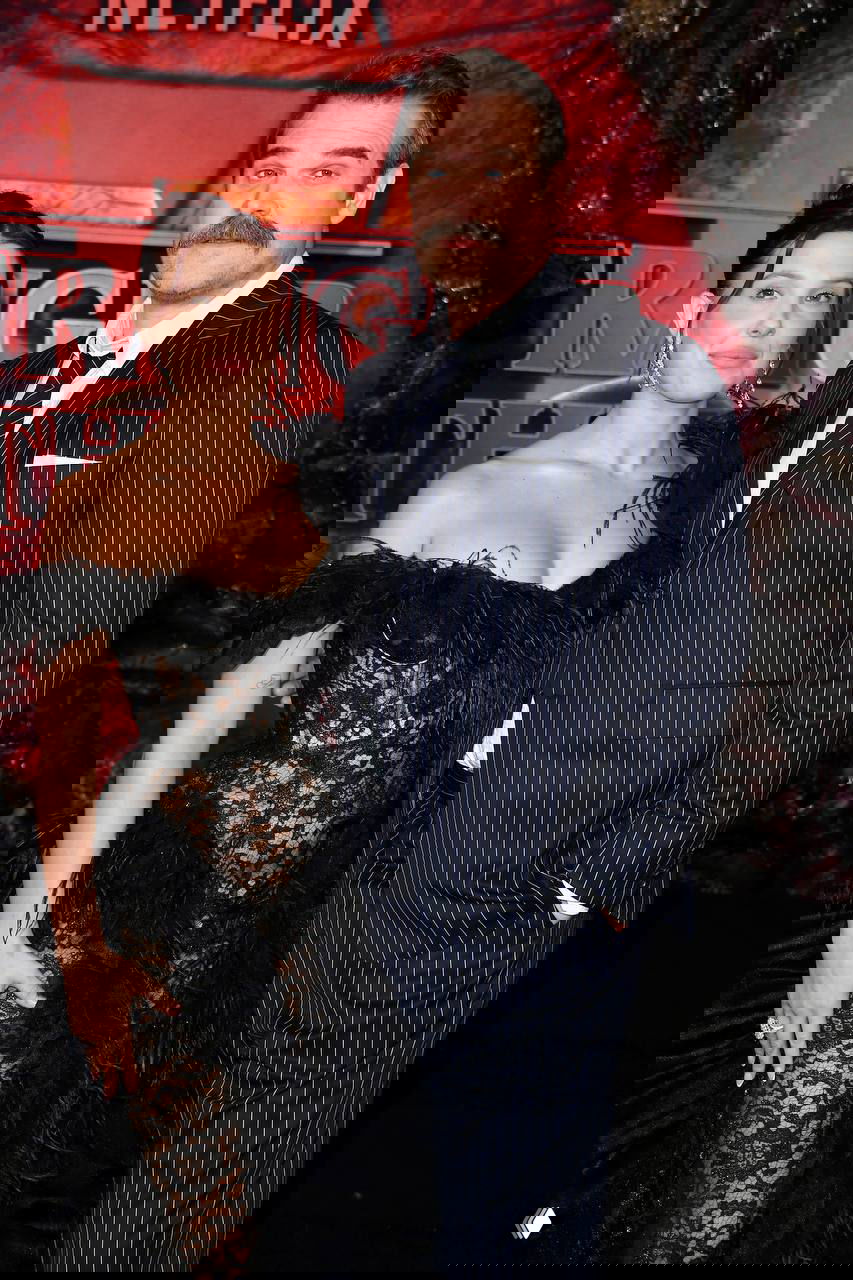 Милли Бобби Браун в черном кружевном платье и Дэвид Харбор в полосатом костюме позируют на красной дорожке премьеры рядом с логотипом Netflix