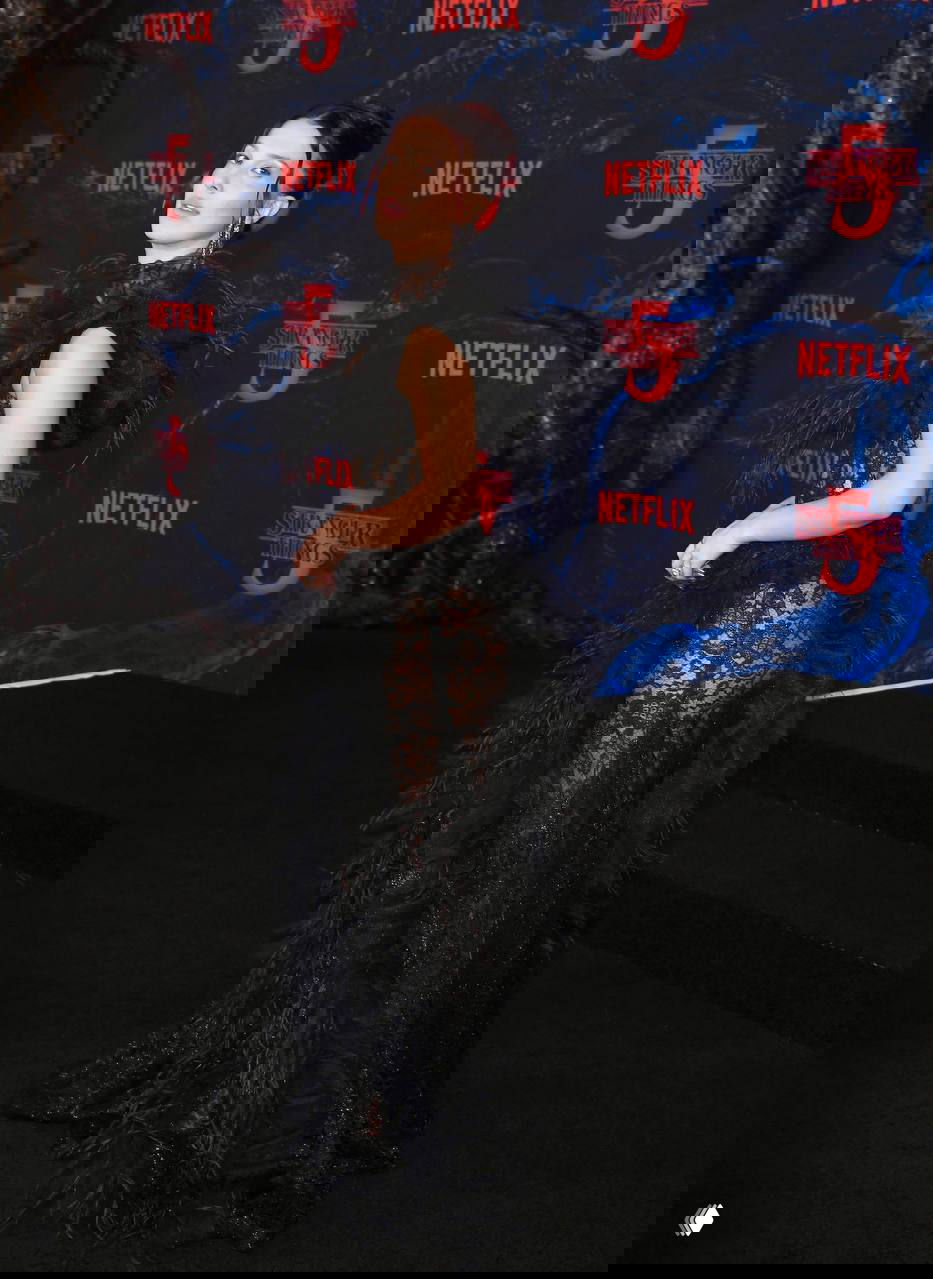 Крупный портрет Милли Бобби Браун в длинном черном кружевном платье на фоне постера и логотипа премьеры сериала на красной дорожке