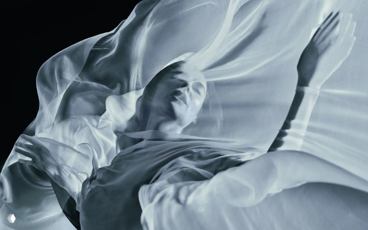Монохромный кадр с полупрозрачными слоями ткани и мягким светом, модель словно плывёт в тканевом облаке — эстетика небес и сна.