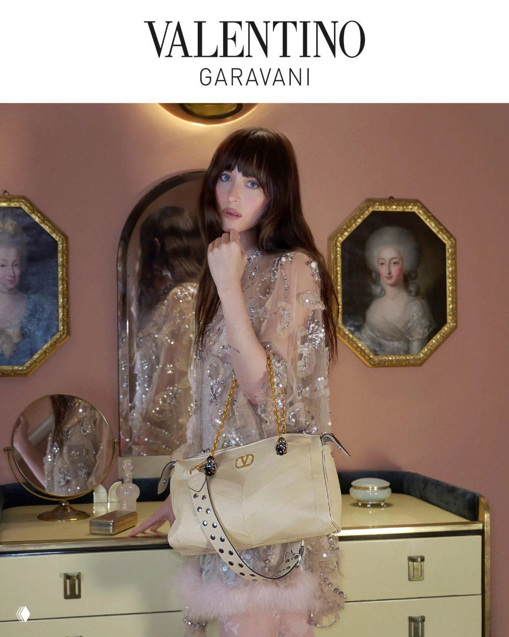 Дакота Джонсон в светлом прозрачном платье держит бежевую сумку Valentino на фоне интерьерной композиции и зеркала — рекламный кадр.