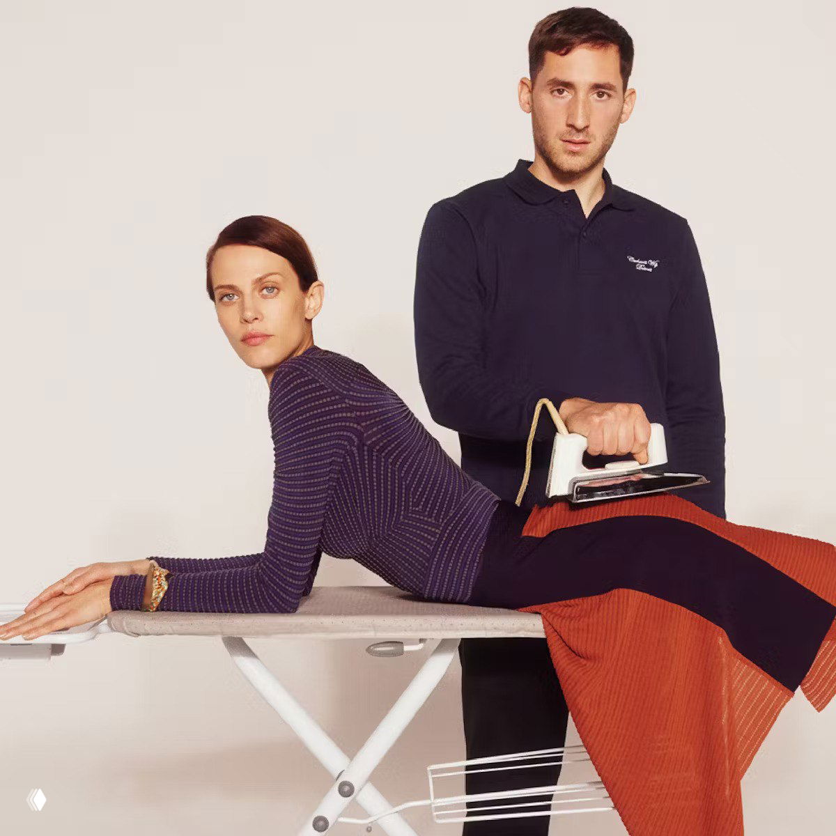 Модная фотография: в концептуальном кадре модель лежит на гладильной доске, рядом мужчина держит утюг в руках, нейтральный фон.