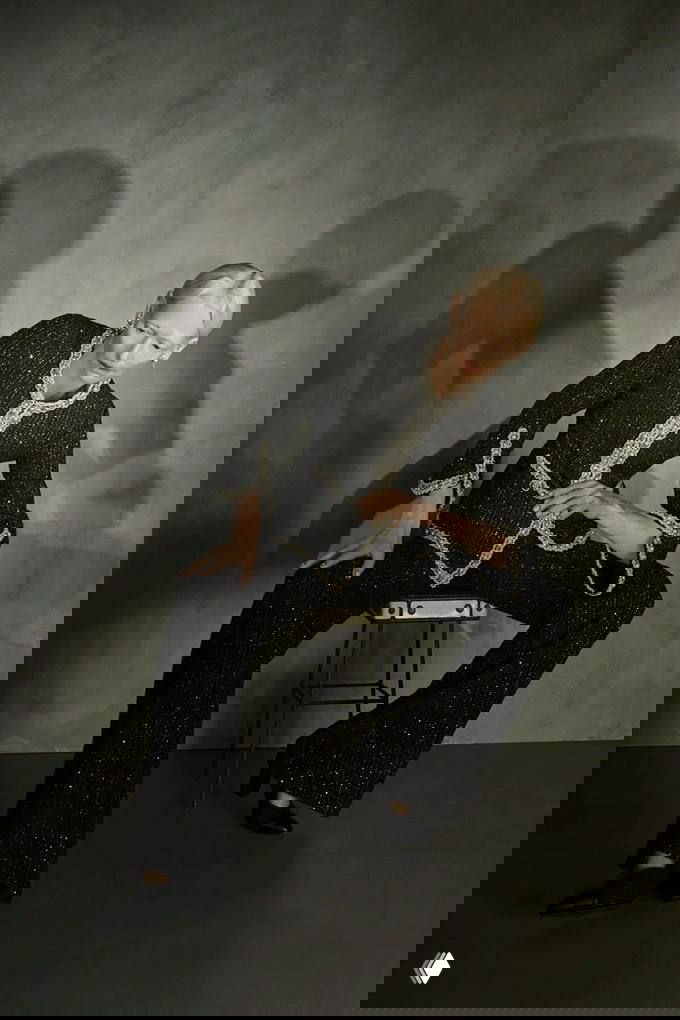 Чёрно‑белое фото: модель сидит на табурете в твидовом жакете и широких брюках Chanel; текстура ткани и контрастная отделка видны в кадре.