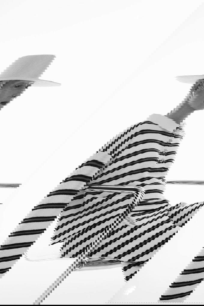 Крупный студийный портрет в профиль: полосатый свитер и брюки, минималистичная шляпа и чистый свет создают сдержанную эстетическую картину.