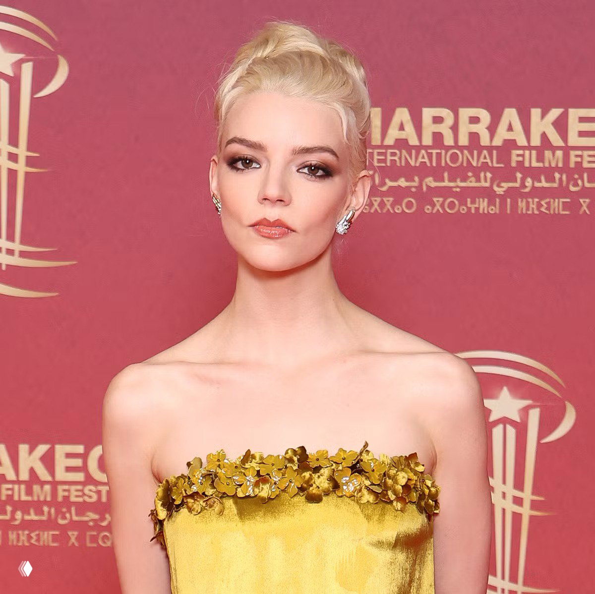 Портрет анфас: Аня Тейлор‑Джой в платье с цветочной отделкой по лифу, минималистичный макияж и высокая причёска на фоне логотипов фестиваля.