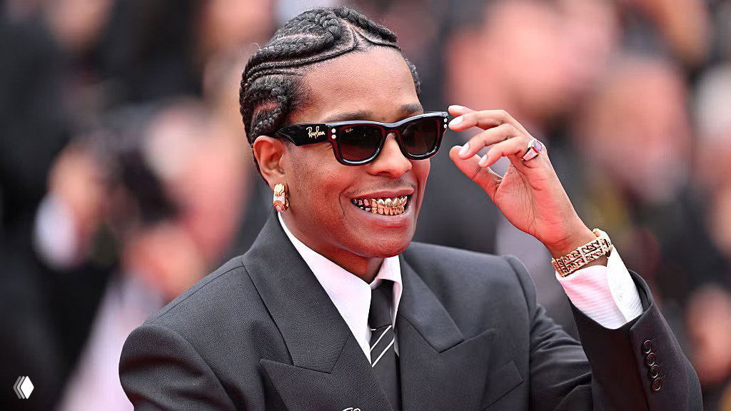 A$AP Rocky в тёмном костюме и солнцезащитных очках на красной дорожке, крупный план: улыбка, золотые украшения и стильный образ.