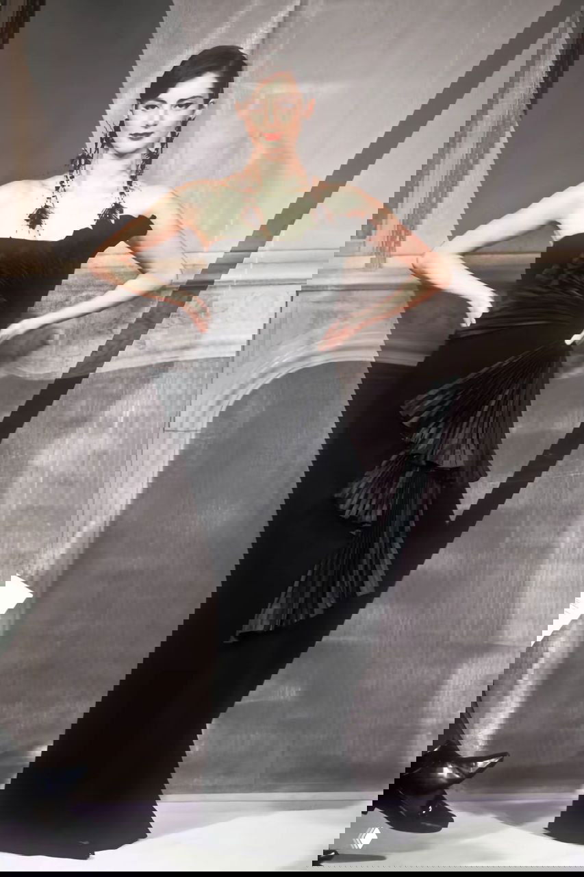 Модель в элегантном чёрном платье с асимметричными плиссировками и скульптурным драпированием на фоне логотипа показа Valentino.