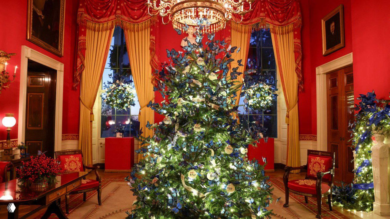 Красная гостиная Белого дома с большою украшенной ёлкой, обилием огней и декоративными элементами, висящая люстра подчёркивает праздничную сцену.