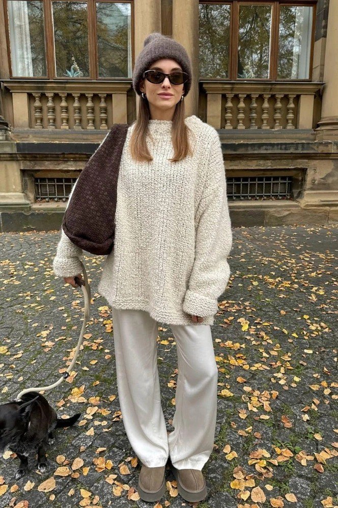 Женщина в крупном светлом свитере и шапке, свободные светлые брюки и сумка через плечо, на прогулке по парку с опавшими листьями — тёплый уютный лук.