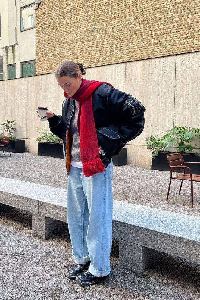 Образ с чёрной бомбер-курткой, ярко-красным шарфом и светлыми джинсами, девушка держит бумажный стакан — повседневный городской комплект для зимы.
