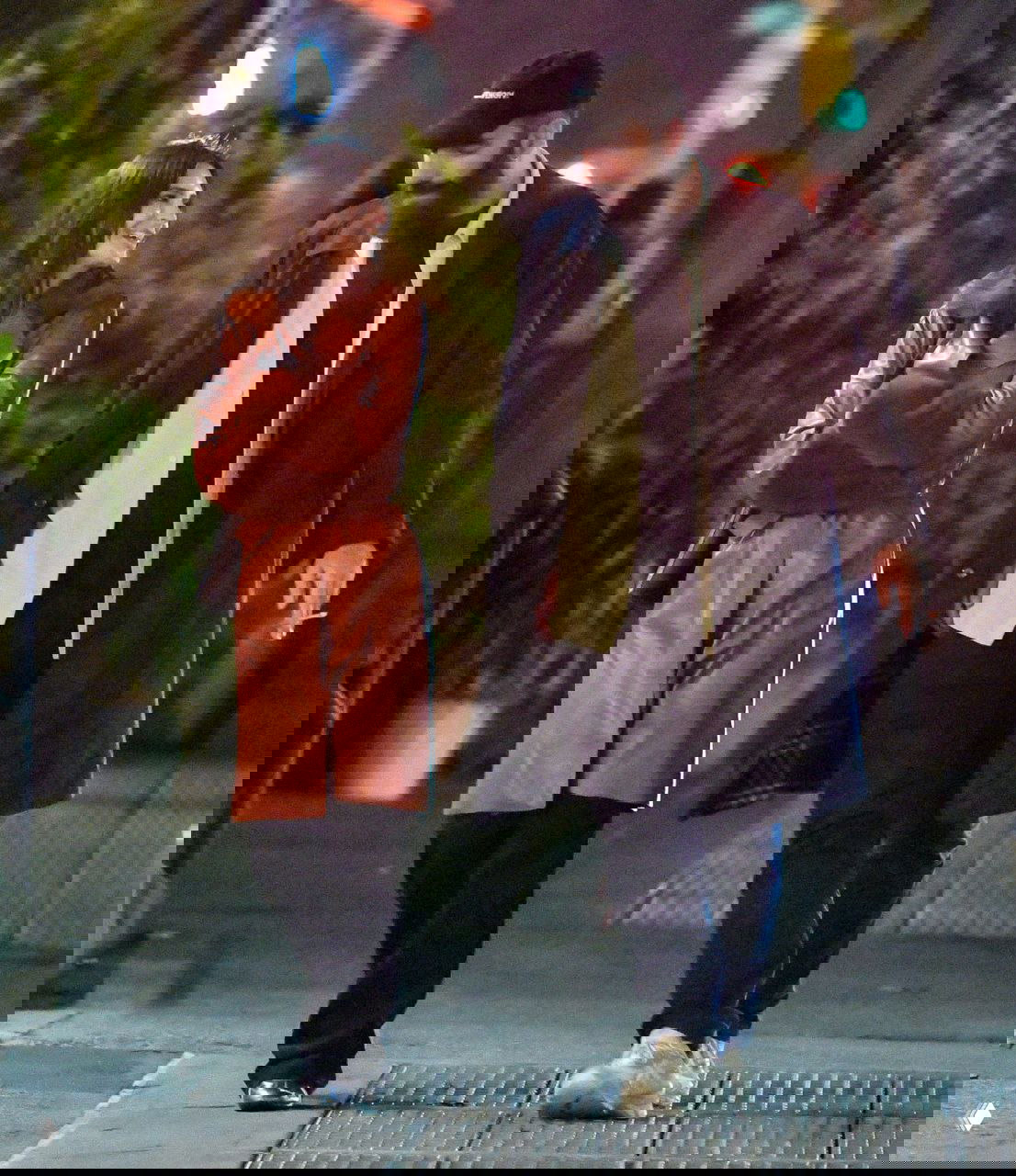 Эмили Ратаковски в коричневом плаще идёт рядом с мужчиной в тёмном пальто и кепке, оба улыбаются, уличная сцена ночью