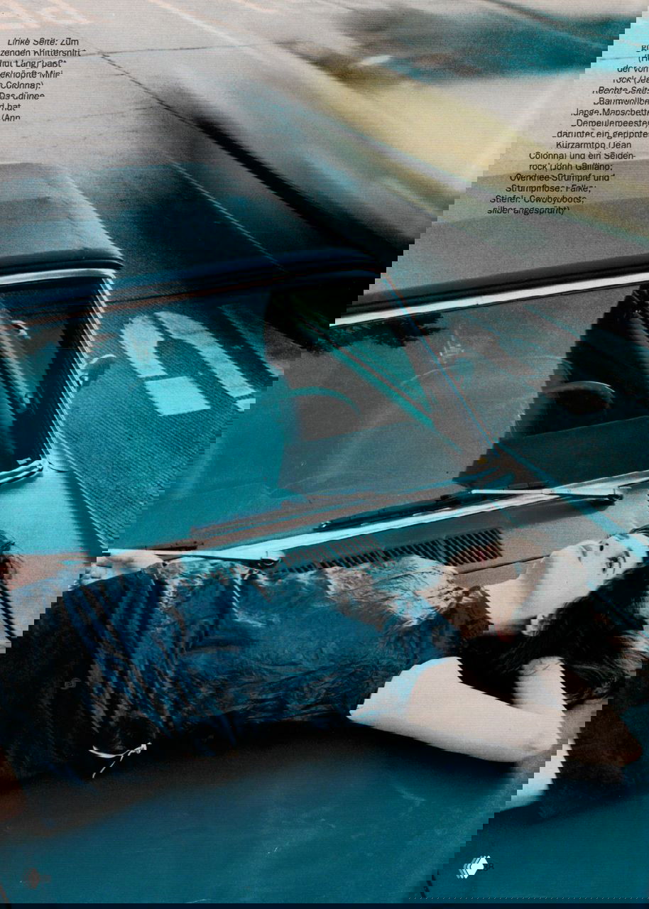 Кадр с автомобильной сценой: модель лежит на капоте старого автомобиля в лаконичном платье и панк‑элементах, съёмка с винтажной зернистостью и прохладной тональностью.