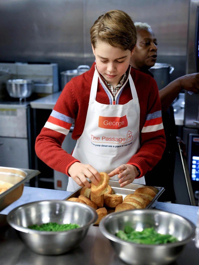 Маленький Джордж в фартуке The Passage раскладывает булочки на противне в кухне приюта, крупный план рук и продуктов, волонтёр рядом.