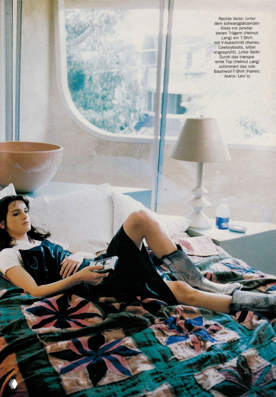 Интерьерный кадр: модель отдыхает на кровати, в ковбойских ботинках и лёгком платье, мягкий дневной свет и фактурное покрывало создают домашнее, неформальное настроение.