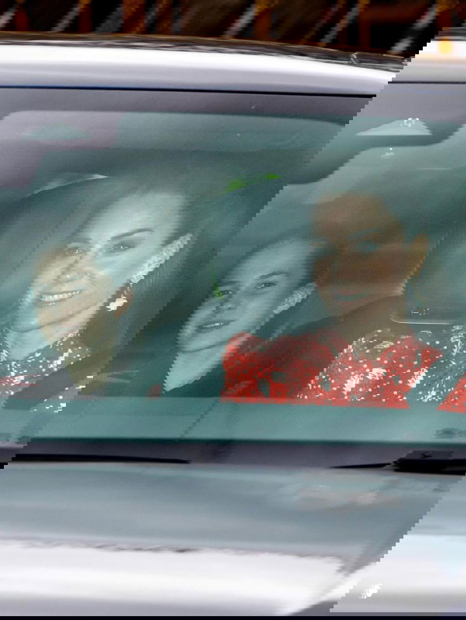 Женщина в ярком красном платье с белым принтом и ребёнок на заднем сиденье автомобиля, кадр снят изнутри через лобовое стекло папарацци.