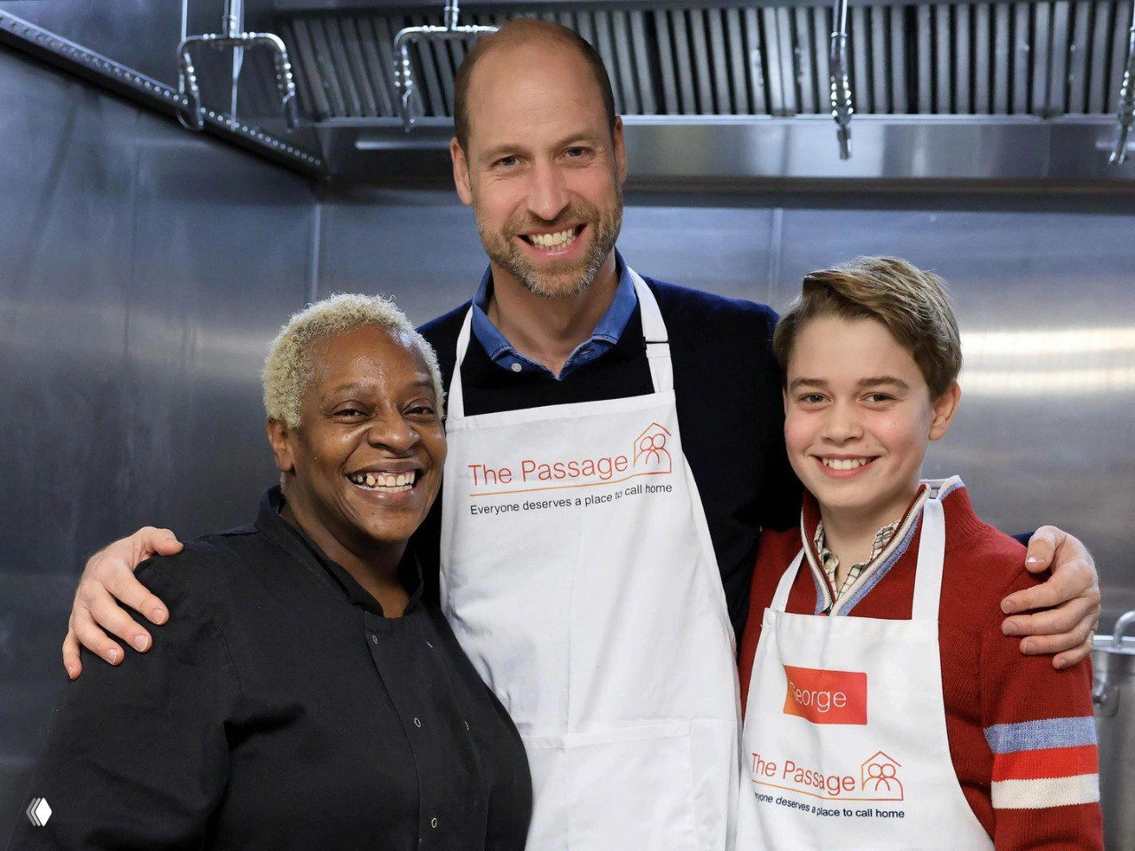 Принц Уильям и Джордж в фартуках The Passage улыбаются рядом с сотрудницей приюта на фоне профессиональной кухни, групповая фотография в дружелюбной атмосфере.