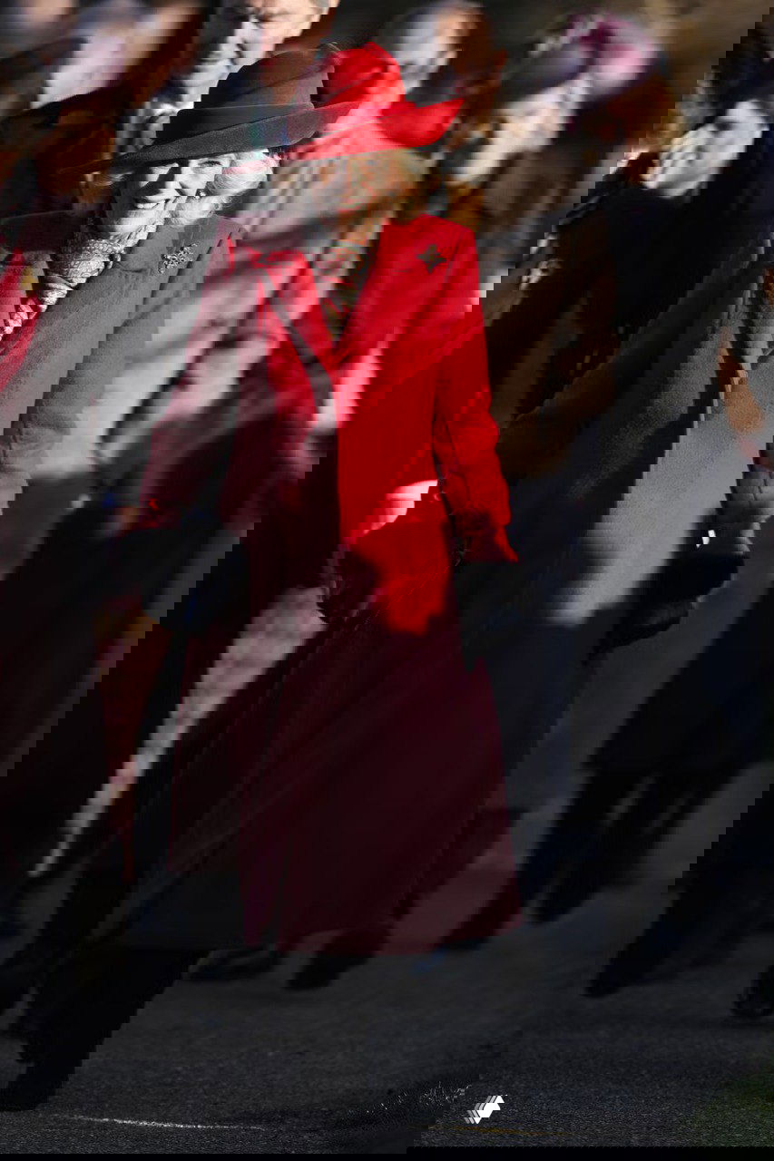 Крупный план женщины в алом пальто и шляпе: видно детали образа — пальто, шляпа и шарф, в глубине — гости службы и праздничная обстановка.