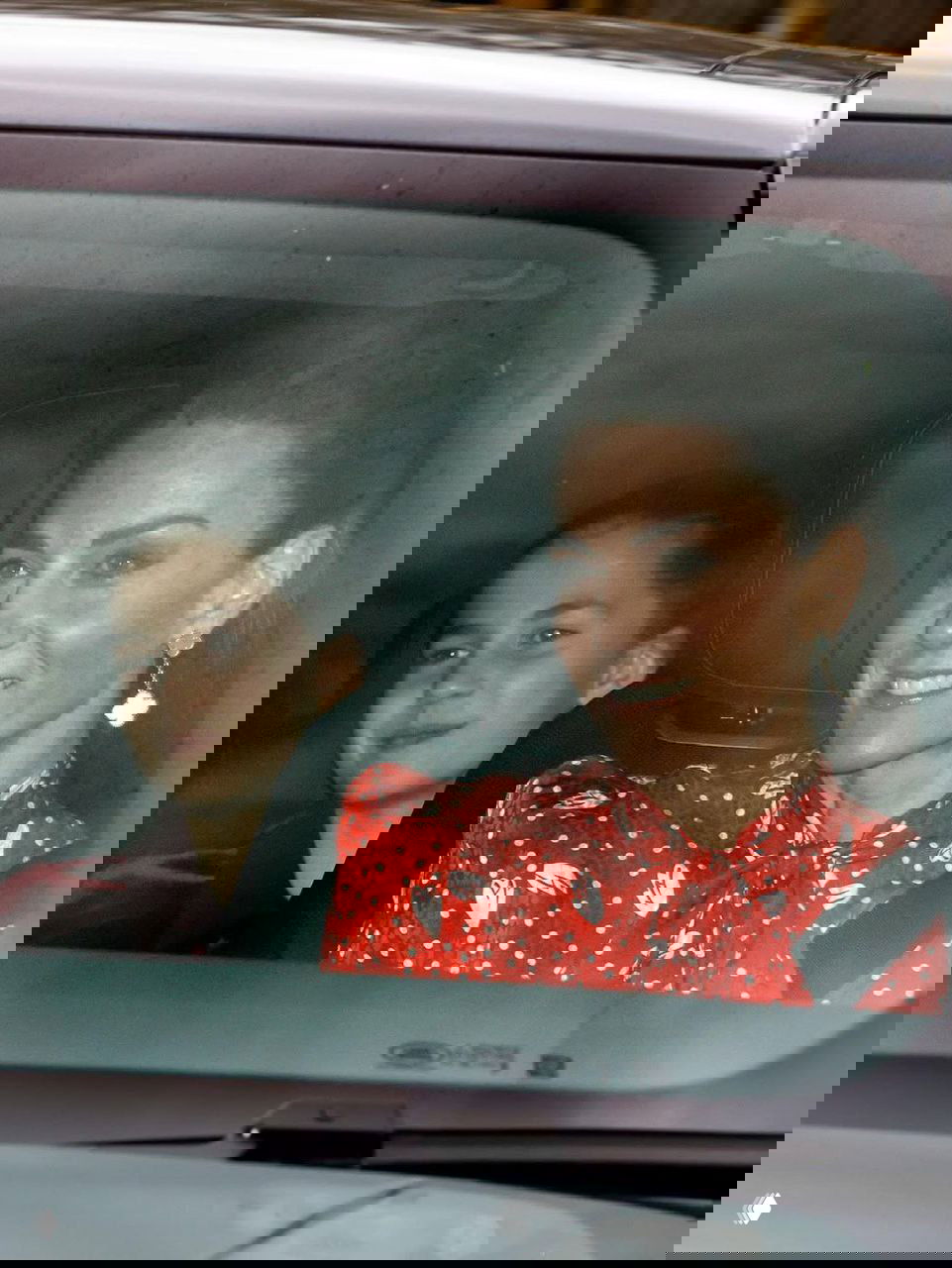 Женщина в красном платье с белым рисунком улыбается в салоне автомобиля, за ней видна юная пассажирка; кадр папарацци изнутри машины.