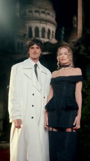 Марго Робби рядом с коллегой у красной дорожки в чёрном мини‑платье Thom Browne, образ в контрасте с плащом спутника и вечерним освещением.