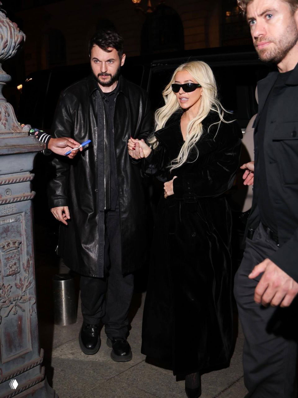 Ночной кадр: женщина в длинном тёмном пальто и солнцезащитных очках выходит из здания вместе с спутником, городской фон, уличное освещение.