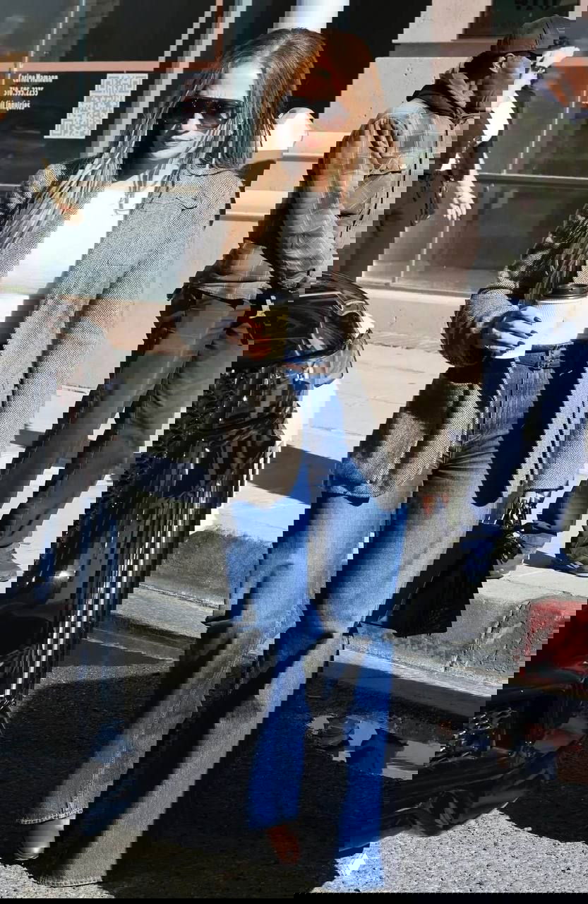 Днёвый кадр: женщина в пиджаке и джинсах держит кофе на тротуаре — повседневный городской стиль с элементами smart-casual.