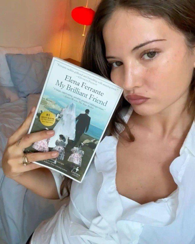 Женщина в белой блузке держит книгу Elena Ferrante «My Brilliant Friend» на кровати — пример поэтичной эстетики и винтажного аксессуара.