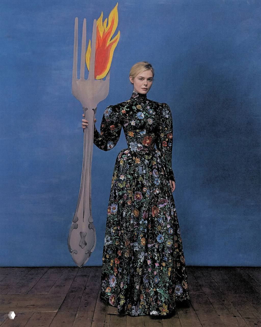 Полный рост Эль Фаннинг в длинном платье с цветочным узором держит крупную декоративную вилку с огнём — театральная постановка.