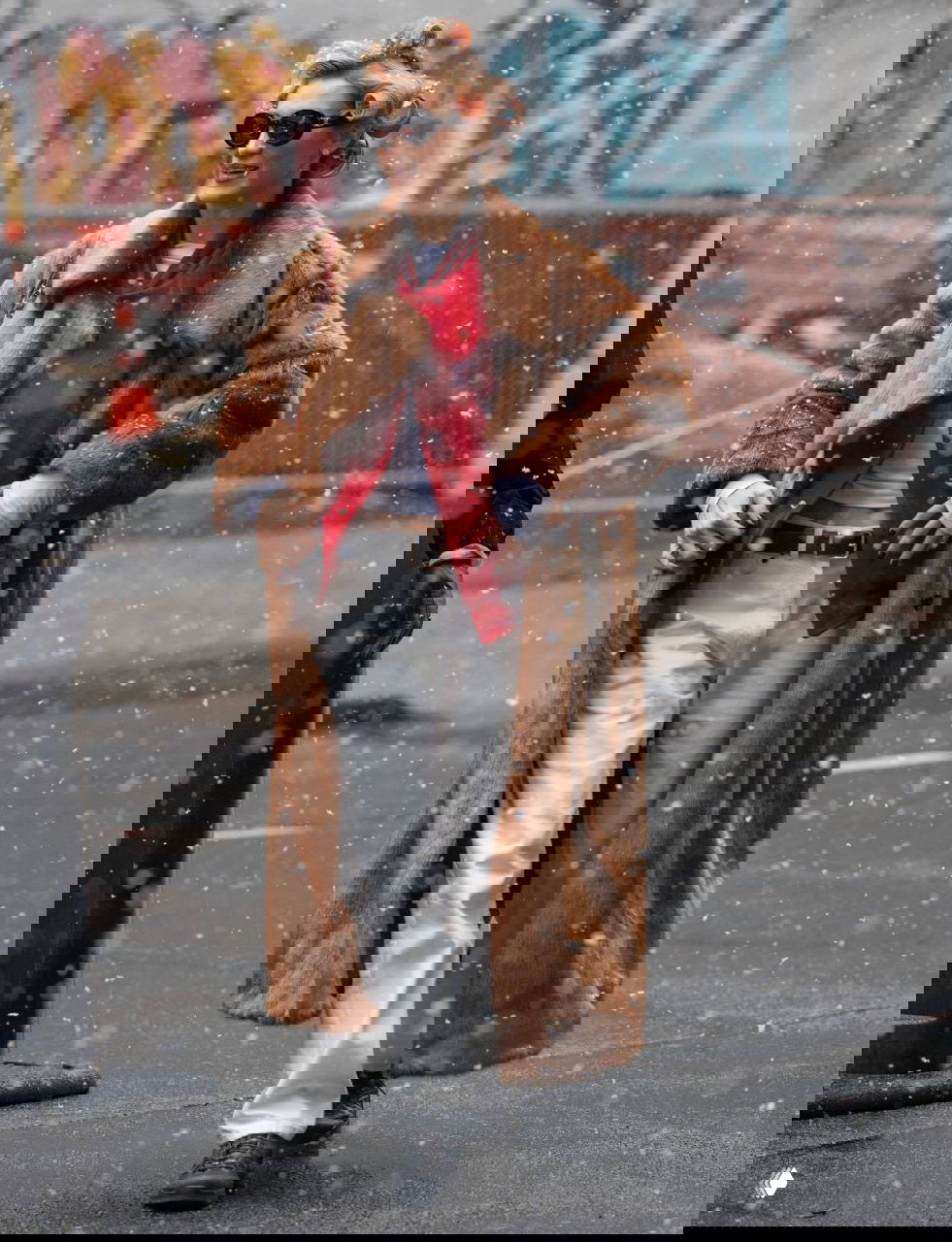 Уличный снимок в снегопад: человек в длинной шубе и стильных очках идёт по заснеженной улице, заметен контраст тёплой верхней одежды и городского фона.