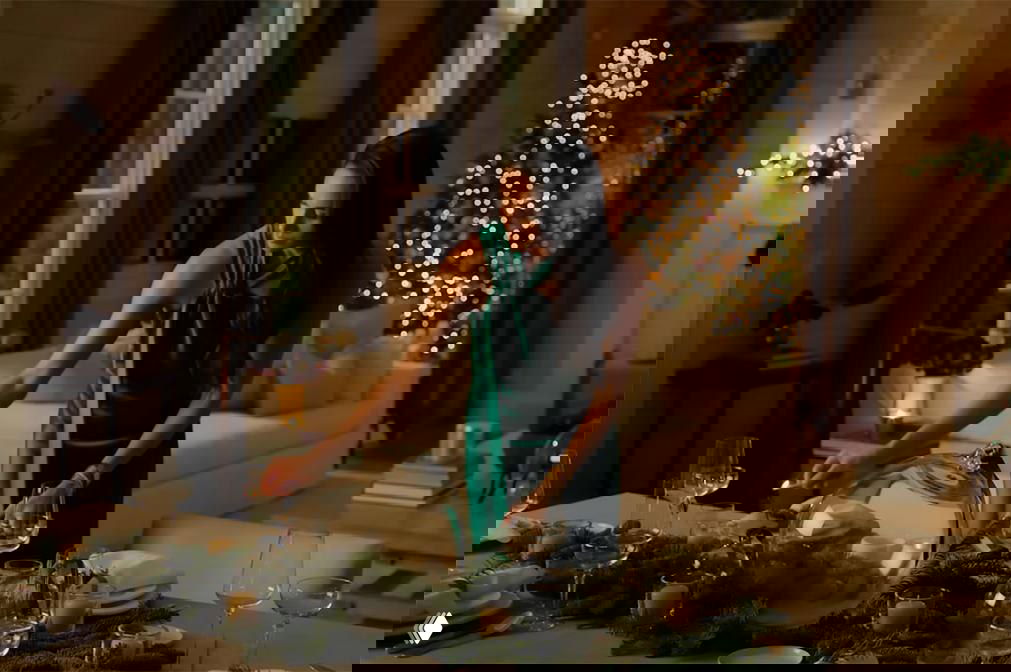 Женщина в изумрудном платье расставляет посуду за праздничным столом в уютной комнате с украшенной рождественской ёлкой на заднем плане.