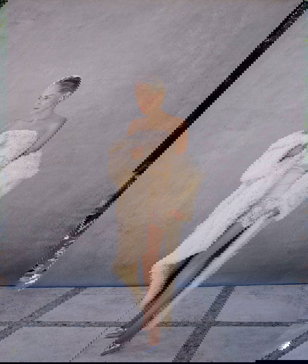 Полнофигурный кадр: женщина в лёгком кружевном платье и длинном кремовом меховом пальто позирует на нейтральном фоне — высокомодный образ.