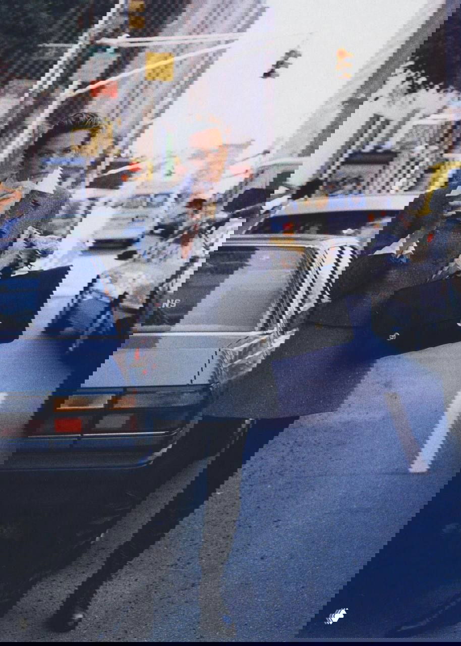 Модель в деловом жакете и мини-юбке держит кофе и портфель на оживлённой городской улице, рабочий городской образ 90-х.