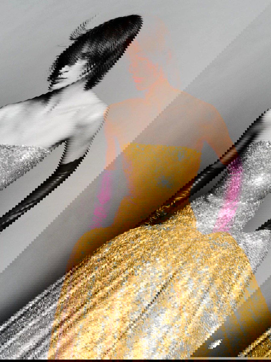 Модель в золотом блестящем платье без бретелей и фиолетовых перчатках позирует на светлом фоне — гламурный вечерний образ бренда.