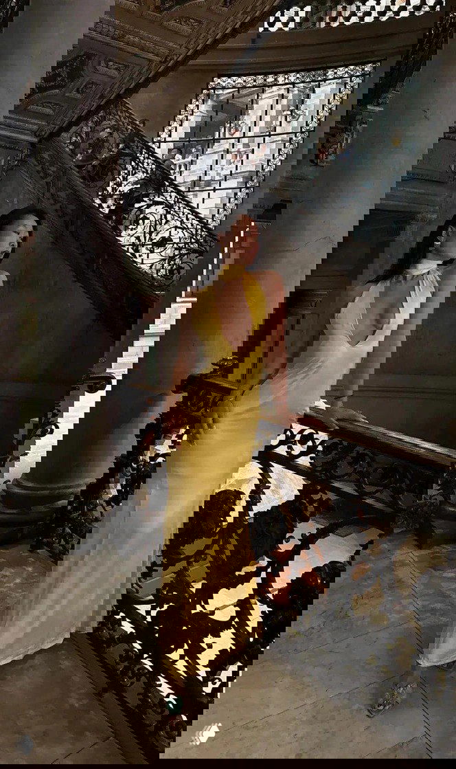 Женщина в длинном жёлтом атласном платье стоит на парадной лестнице старинного интерьера; образ с элегантной обувью и минимальными украшениями.