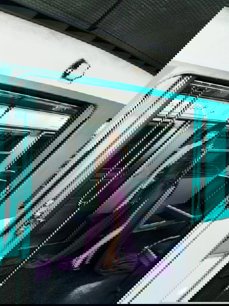 Модель в длинном фиолетовом платье с драпировкой стоит в вагоне метро у поручня — неожиданное сочетание высокой моды и городского пейзажа.