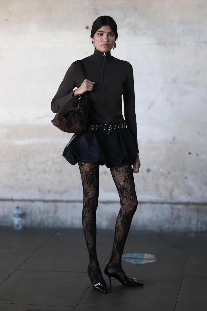 Модель в чёрном мини‑юбочном образе и кружевных колготках на фоне урбанистической сцены — сочетание готики и романтики в моде.