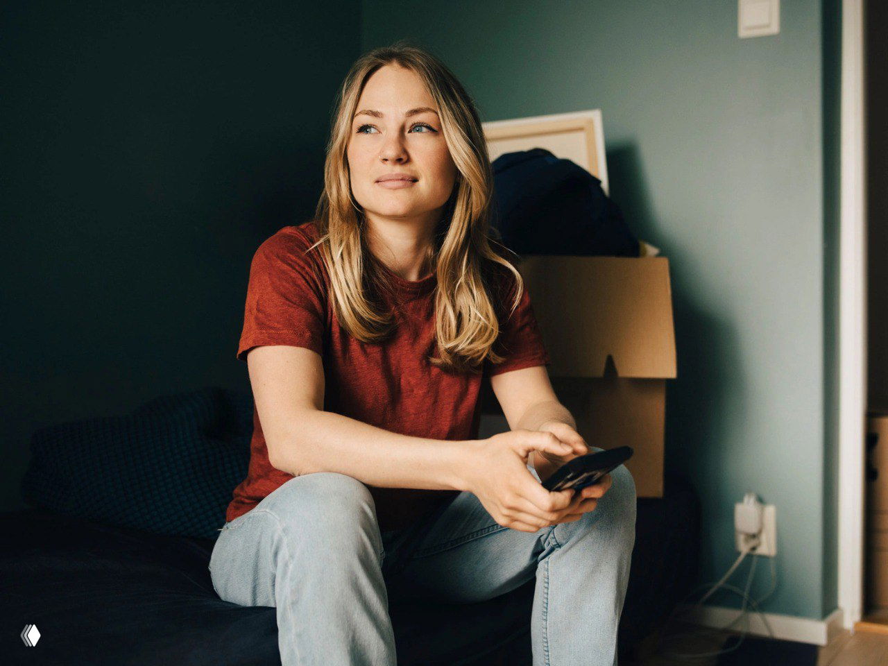 Молодая женщина в комнате сидит с телефоном в руках и задумчиво смотрит в сторону — визуал к материалу о подведении итогов и планах.