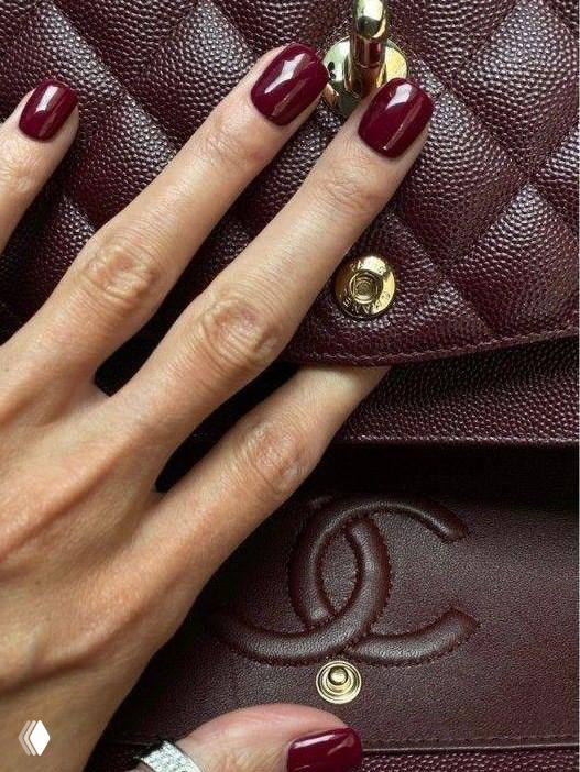 Крупный план руки с тёмно-вишнёвым маникюром, пальцы опираются на бордовую лакированную сумку с тиснением логотипа, элегантный образ.