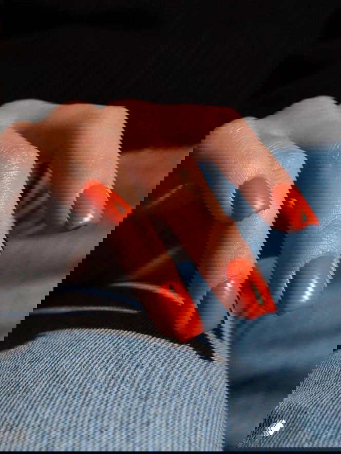 Матовый ярко-оранжевый маникюр на квадратных ногтях, рука расположена на джинсовой ткани, модный смелый оттенок для праздника.