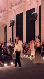 Модель в белом жакете и чёрных брюках на фоне торжественной архитектуры зала; контраст строгого кроя и кутюрных деталей.