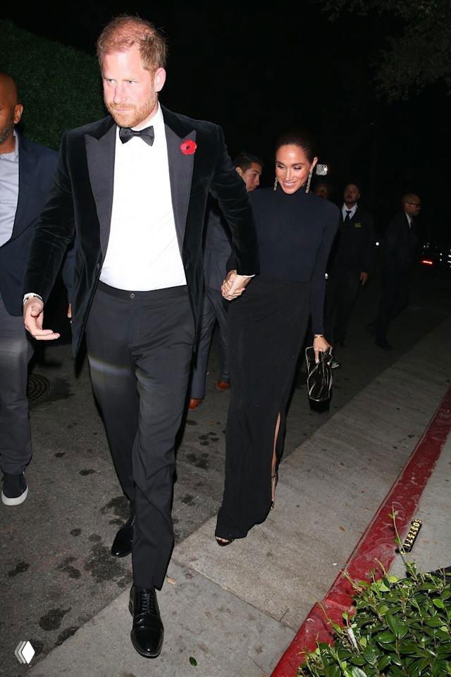 Пара идёт по тротуару вечером: мужчина в смокинге и женщина в длинном чёрном платье, фото сделано папарацци при выходе с мероприятия.