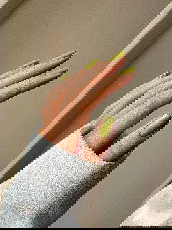 Рука с салатово-зелёным маникюром на аккуратных квадратных кончиках ногтей, светлый нейтральный фон подчёркивает свежесть оттенка.