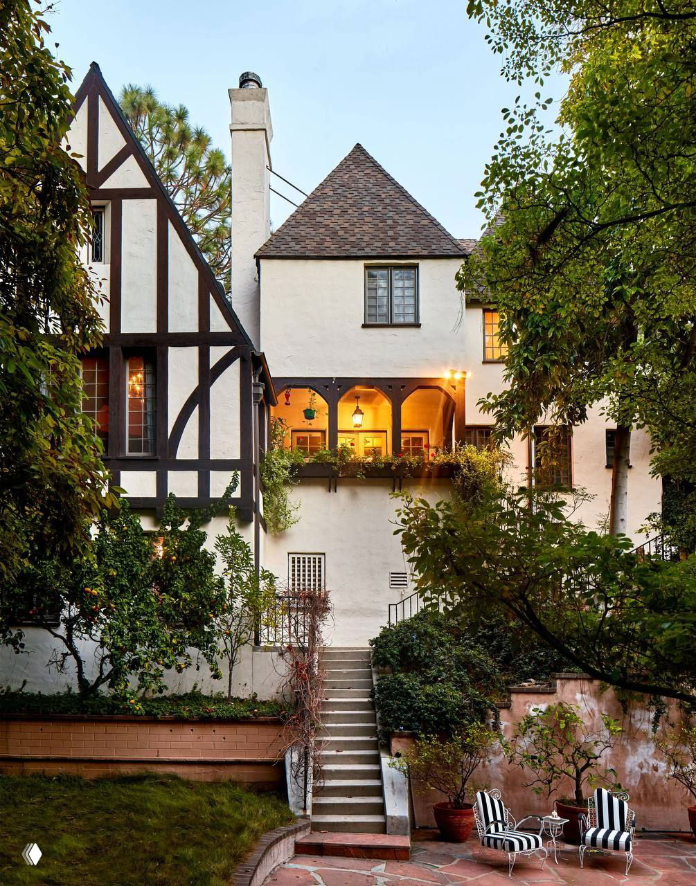 Фасад особняка в стиле Tudor Revival с крыльцом и лестницей, окружённый зелёным садом и патио с креслами — вид со стороны двора.