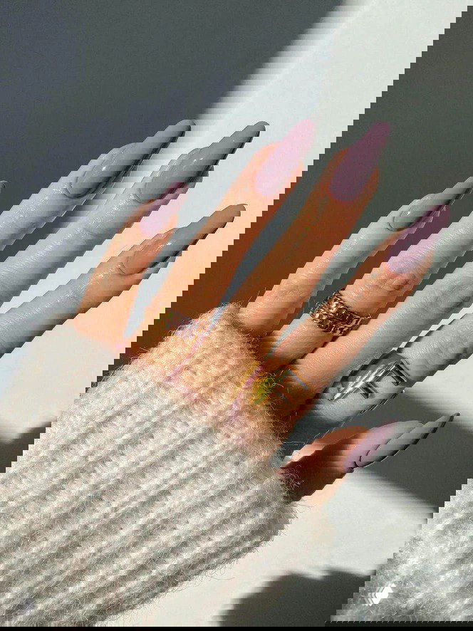 Рука с пастельно-лиловым маникюром и золотыми кольцами на пальцах, мягкий свет и вязаный рукав создают уютный стильный образ.