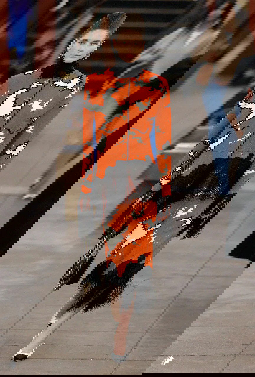 Модель в оранжево‑чёрном комплекте с бахромой и графичным принтом проходит по подиуму — центр внимания на контрастном костюме Chanel.