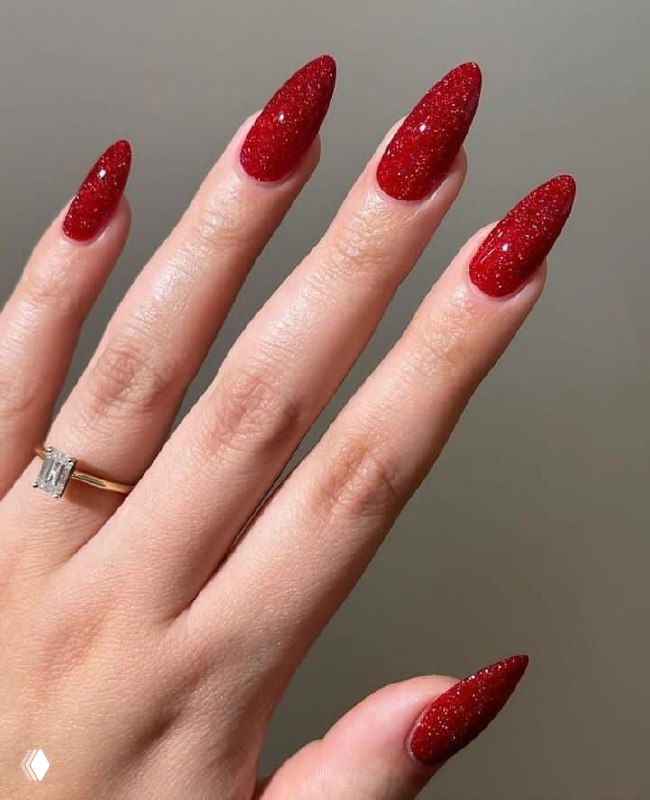 Рука с миндалевидными ногтями в красном глиттере: яркие блёстки, ровная форма и нейтральный серый фон для контраста.