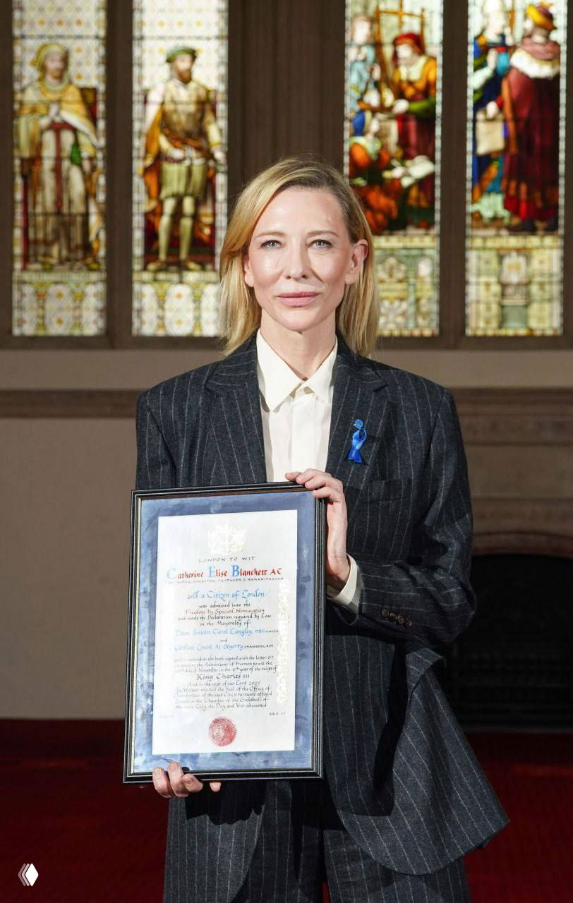 Кейт Бланшетт держит рамку с грамотой на фоне витражного окна в торжественном зале; строгий костюм и эмблема награды заметны.