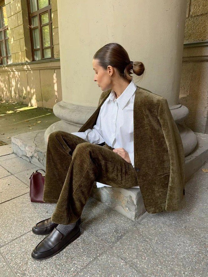 Женщина в коричневом бархатном брючном костюме и лоферах сидит у колонны театрального здания, рядом видно бордовый клатч и тротуар.