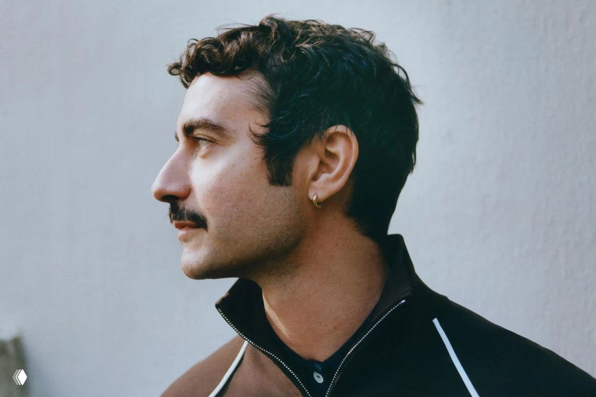 Портрет мужчины в профиль на нейтральном фоне: короткие тёмные волосы, ухо с серёжкой и спортивная куртка — иллюстрация модной новости.