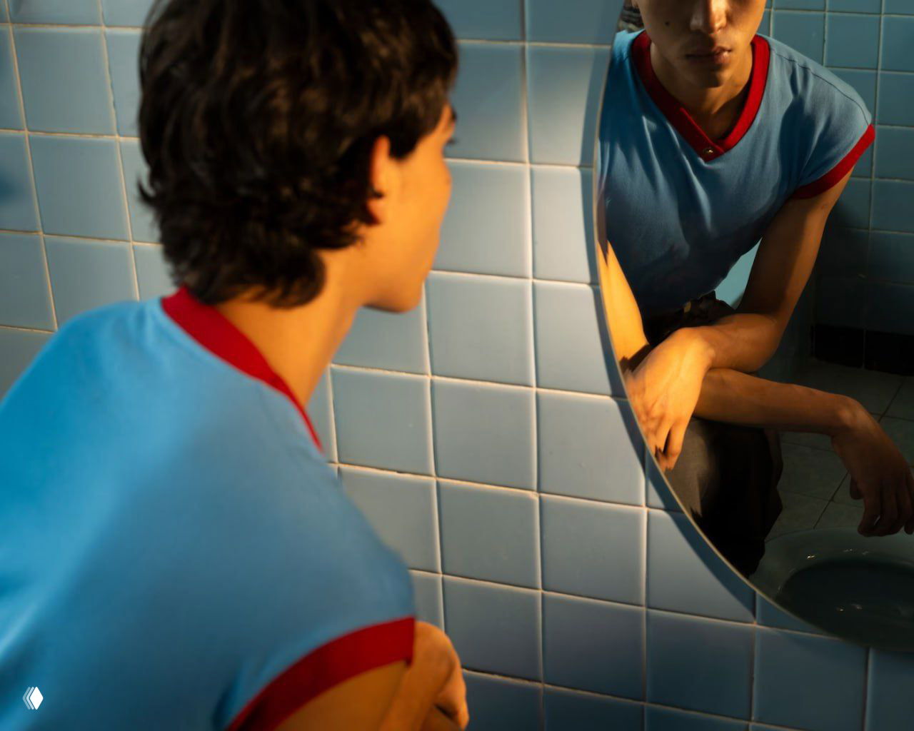 Крупный план в ванной: молодой мужчина смотрит в круглое зеркало на фоне голубой плитки, приглушённый свет и рефлексы стекла.