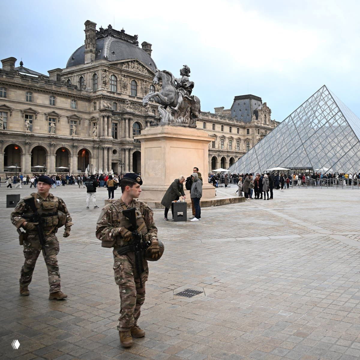 Двор Лувра с пирамидой и фасадом музея; на переднем плане двое вооружённых солдат патрулируют площадь, вокруг видны посетители и очередь.